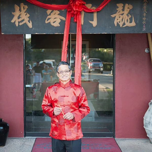 A smiling man in a red traditional Chinese jacket stands in front of a restaurant entrance.