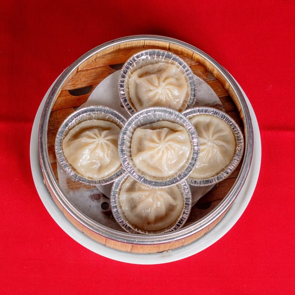 Six soup dumplings in foil cups arranged in a bamboo steamer on a red tablecloth.