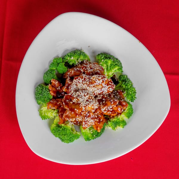 Sesame chicken with broccoli florets on a white plate, set against a vibrant red background.