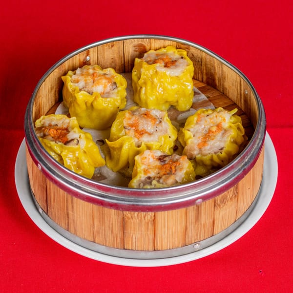 Six yellow dim sum dumplings with orange topping in a bamboo steamer on a red tablecloth.
