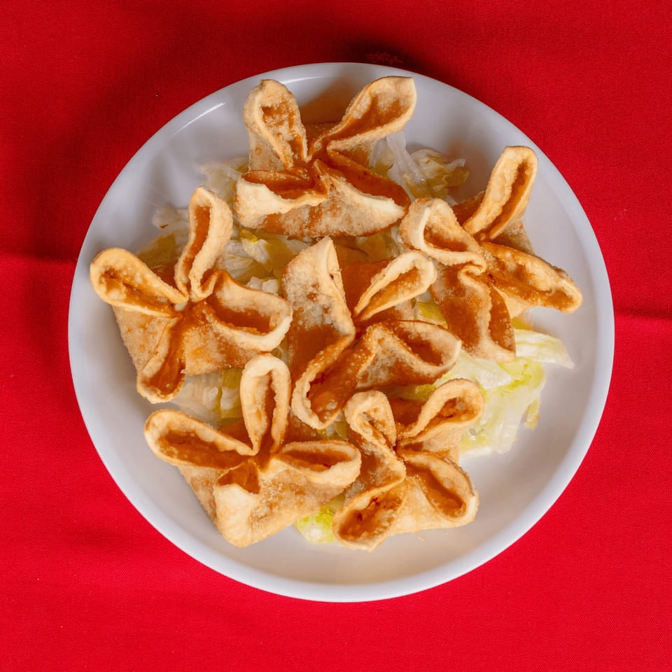 A white plate of golden-brown crab rangoon appetizers on a bed of shredded lettuce, against a red tablecloth.