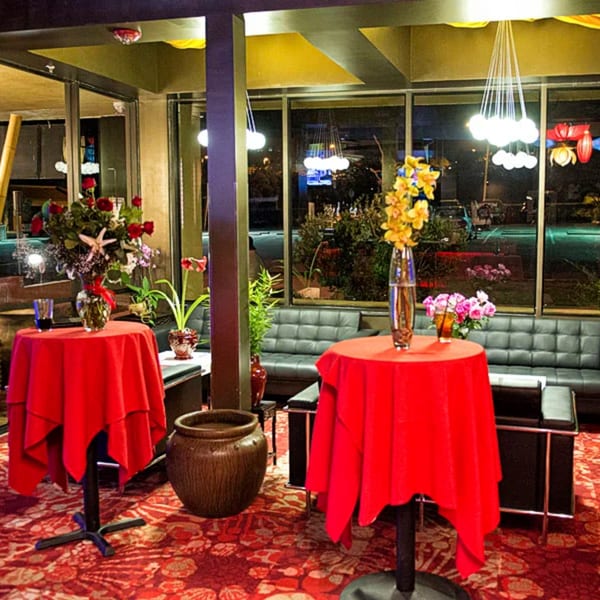 Restaurant interior with red tablecloths, floral arrangements, black couches, and large windows at night.