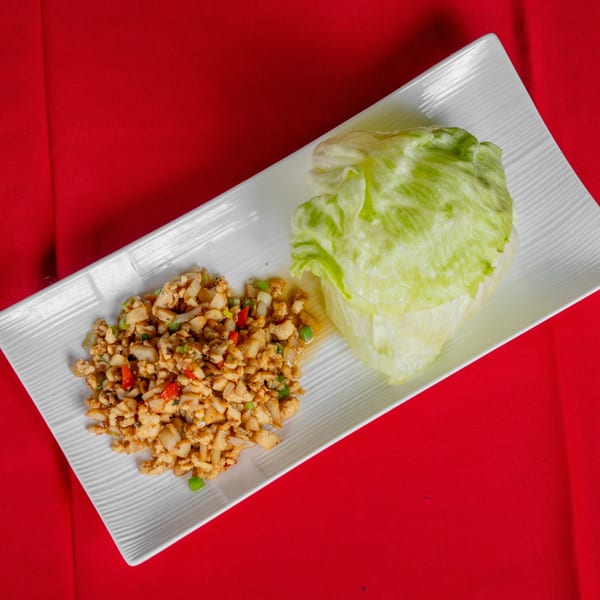 A white rectangular plate with chicken lettuce wraps and a head of lettuce on a red tablecloth.